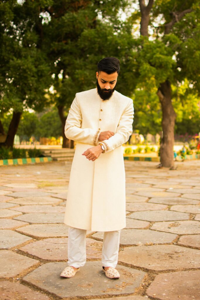 Stylish Indian man in an elegant achkan, adjusting his cufflinks outdoors on a sunny day.